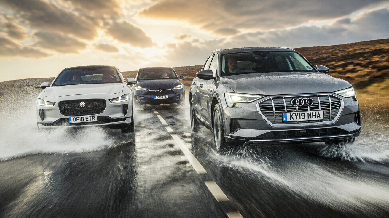 Drei Elektro-SUVs (Audi, Jaguar, Tesla) fahren auf nasser Straße bei bewölktem Himmel.