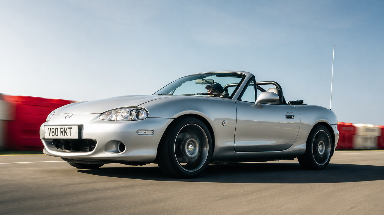 Silberner Mazda MX-5 Cabrio fährt auf sonniger Straße, Fahrer mit Sonnenbrille am Steuer.
