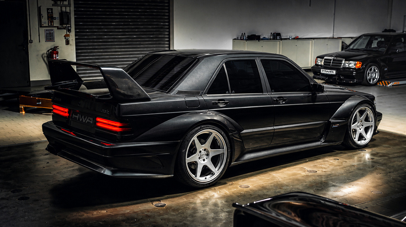 Schwarzes getuntes Sportcoupé mit großem Heckspoiler in beleuchteter Halle, im Hintergrund ein weiterer schwarzer Wagen.