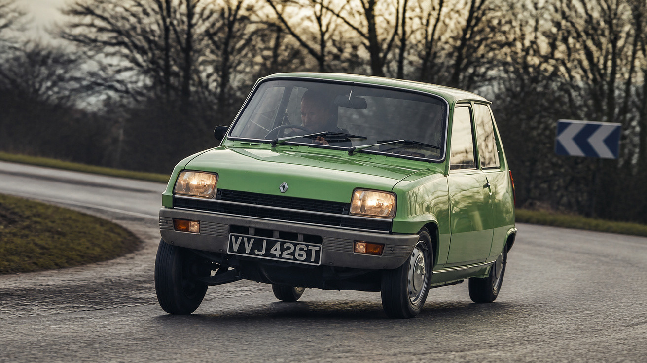 Grüner Renault Oldtimer fährt auf kurviger Landstraße bei bewölktem Himmel und kahlen Bäumen im Hintergrund.