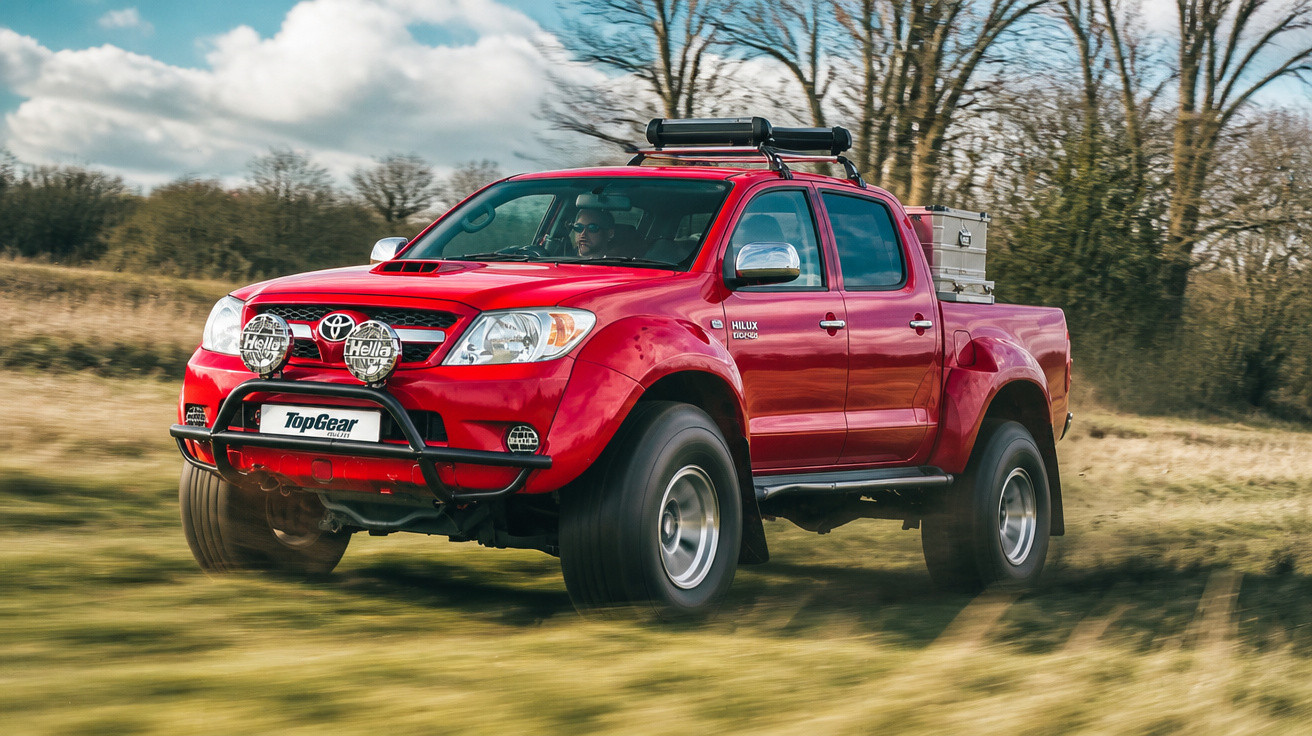 Roter Toyota Hilux Pickup fährt schnell über eine Wiese, mit Offroad-Licht und Dachträger.