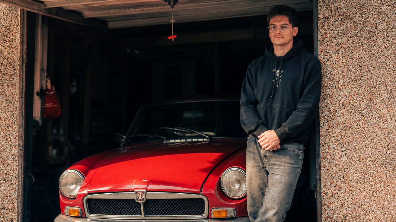 Junger Mann lehnt an der Mauer neben einem roten Oldtimer in einer Garage.