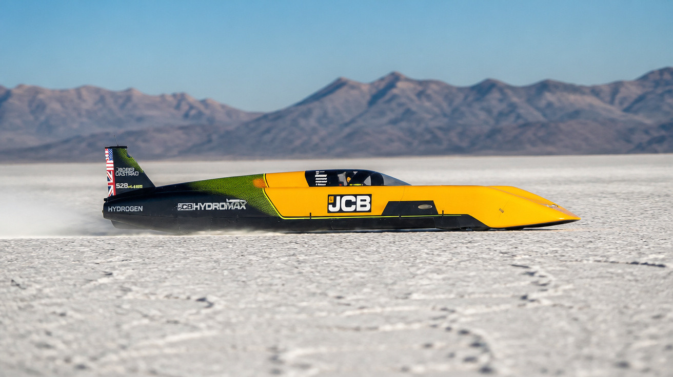 Gelb-schwarzes Landgeschwindigkeitsfahrzeug JCB HYDROMAX fährt über trockenen Salzsee vor Bergkette.