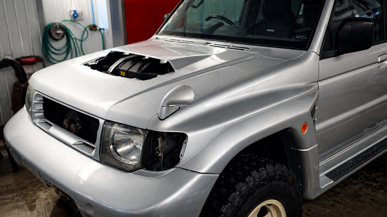 Silbernes Geländewagen-Fahrzeug ohne rechte Frontscheinwerfer in Werkstatt mit offenem Motorhauben-Auslass.