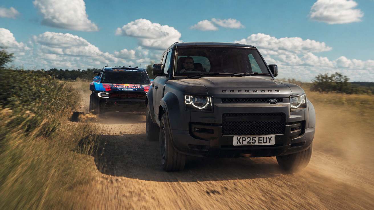 Zwei Geländewagen fahren auf staubigem Feldweg, Vordergrund: schwarzer Land Rover Defender, Hintergrund: Rallye-Fahrzeug.
