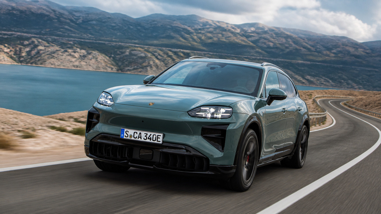 Grüner Porsche SUV fährt auf kurviger Straße entlang eines Sees vor bergiger Kulisse bei bewölktem Himmel.