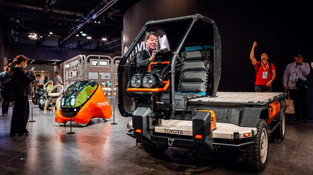 Mann sitzt in einem futuristischen Toyota-Geländefahrzeug auf einer Messe, im Hintergrund weitere Fahrzeuge und Besucher.
