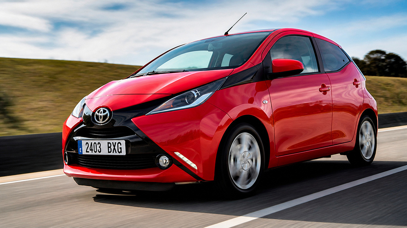 Roter Toyota Aygo fährt auf Straße, Landschaft und Himmel im Hintergrund, Front-3/4-Ansicht.