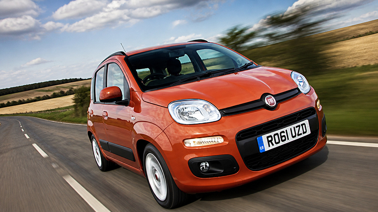 Oranger Fiat Kleinwagen fährt auf Landstraße, unscharfer Hintergrund mit Feldern und Wolken.