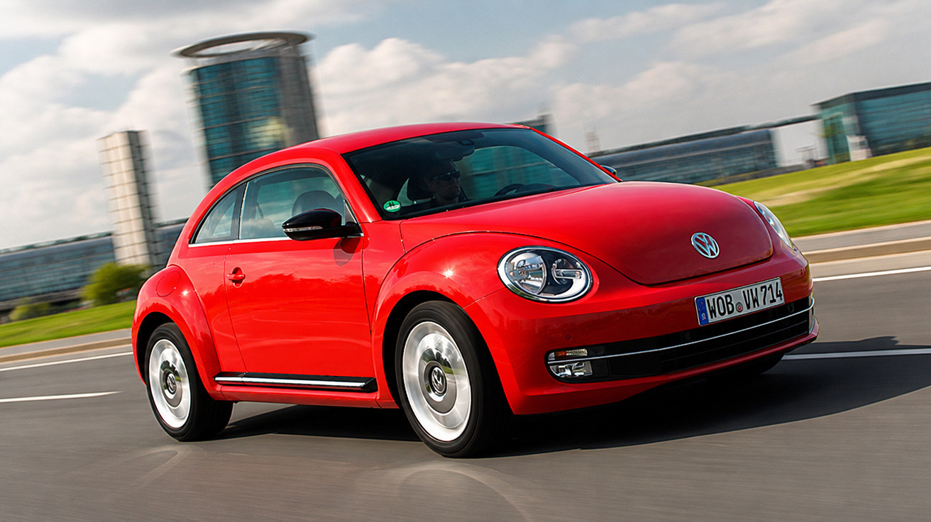 Rotes Volkswagen Beetle Coupé fährt auf Straße vor moderner Stadtlandschaft bei Tageslicht.