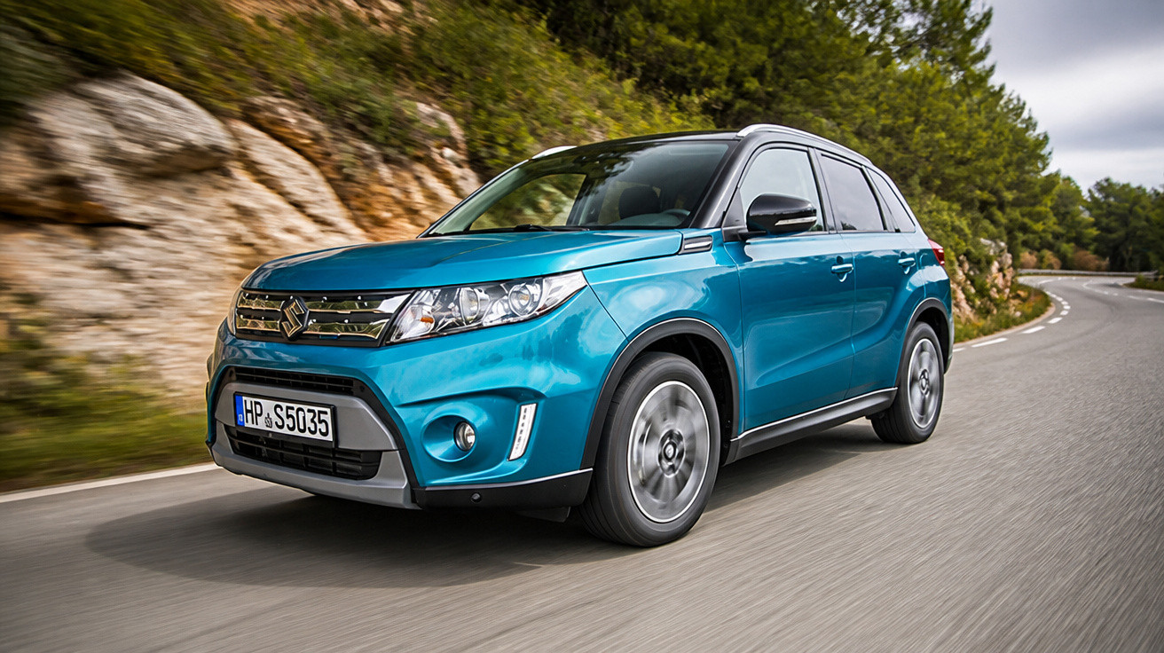 Blauer Suzuki SUV fährt auf einer kurvigen Landstraße neben Felsen und Wald bei bewölktem Himmel.