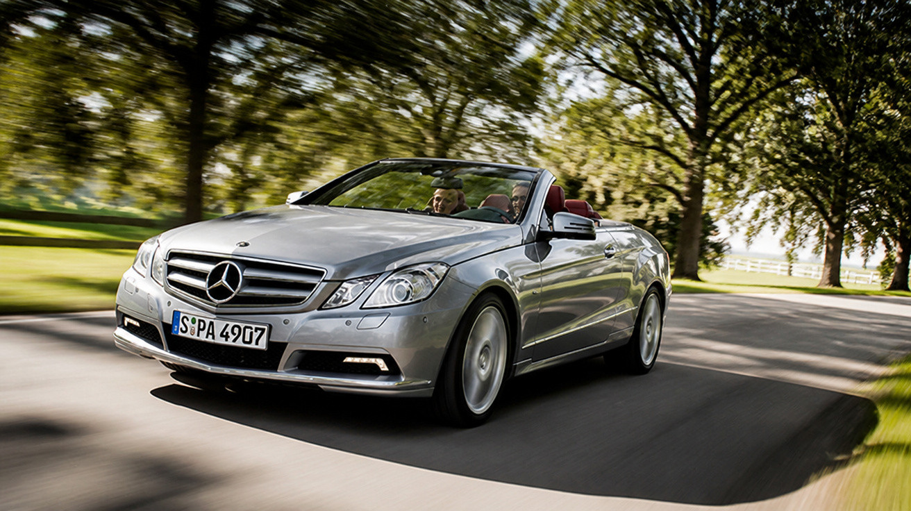 Silberner Mercedes-Benz Cabrio fährt auf einer baumbestandenen Landstraße bei Sonnenschein.