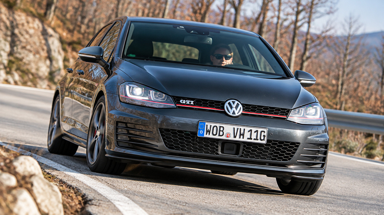 Schwarzer Volkswagen Golf GTI fährt kurvig bergauf auf Asphaltstraße, Fahrer mit Sonnenbrille sichtbar.